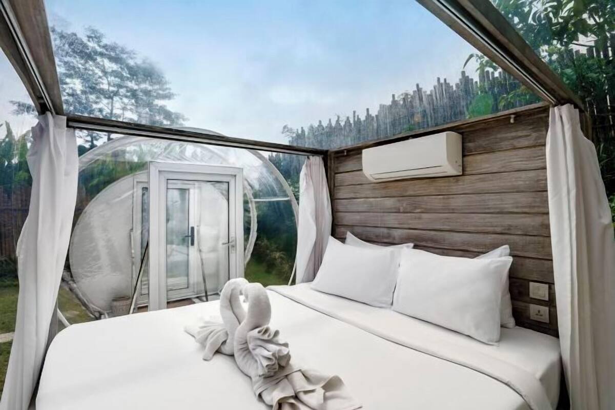 Une chambre à coucher avec un lit, des oreillers blancs, une tête de lit en bois et une vue sur un jardin, visible à travers une fenêtre en verre.