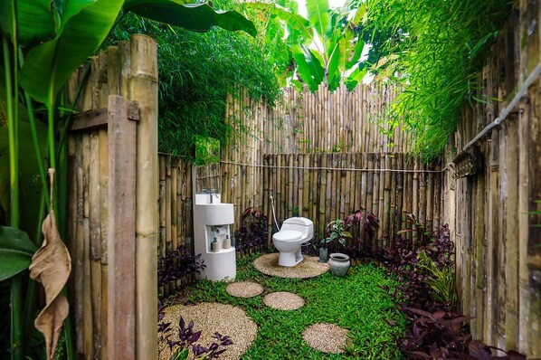 Baño con paredes de bambú, que incluye inodoro, lavabo y zona de ducha, rodeado de abundante vegetación y guijarros.