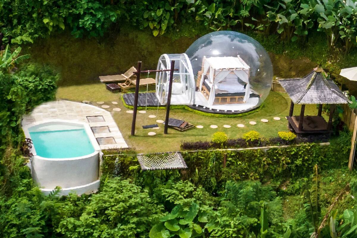 Une maison sphérique en verre, avec un lit à l’intérieur, entourée d’un jardin circulaire agrémenté de pierres plates servant de pas et d’une piscine.