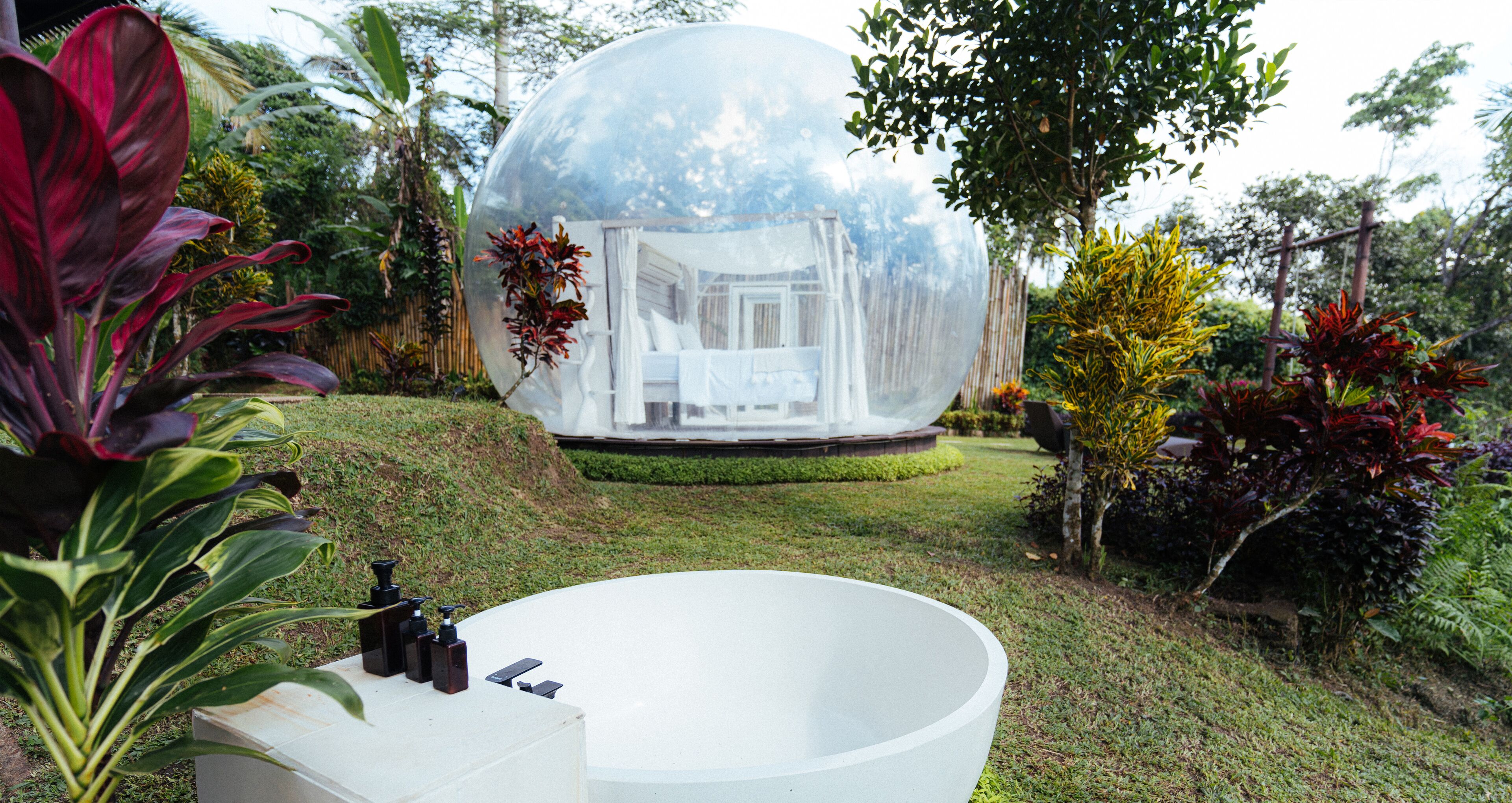 Estructura esférica transparente con interior visible, rodeada de vegetación exuberante y una bañera blanca en primer plano.