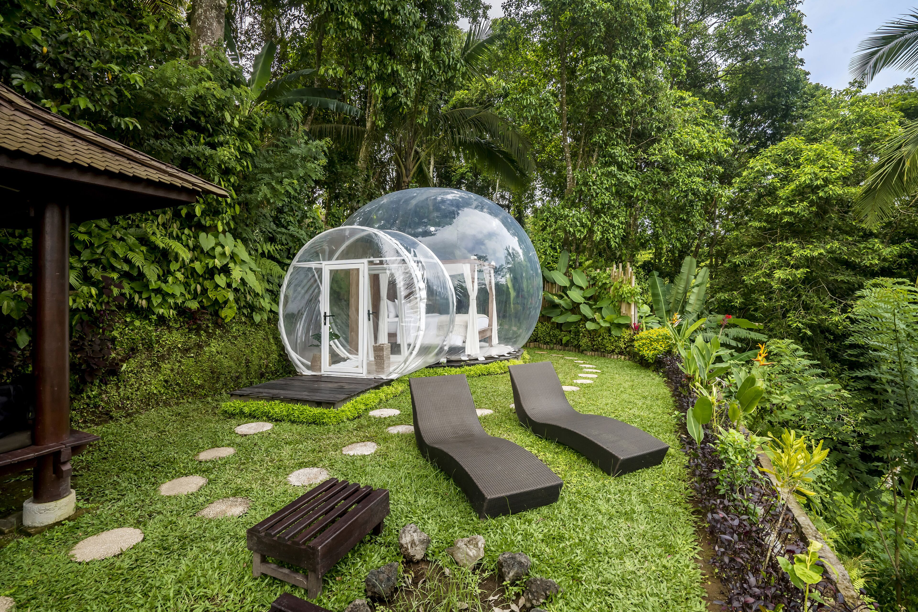A transparent dome structure in a lush garden with a wooden bench and a small gazebo.