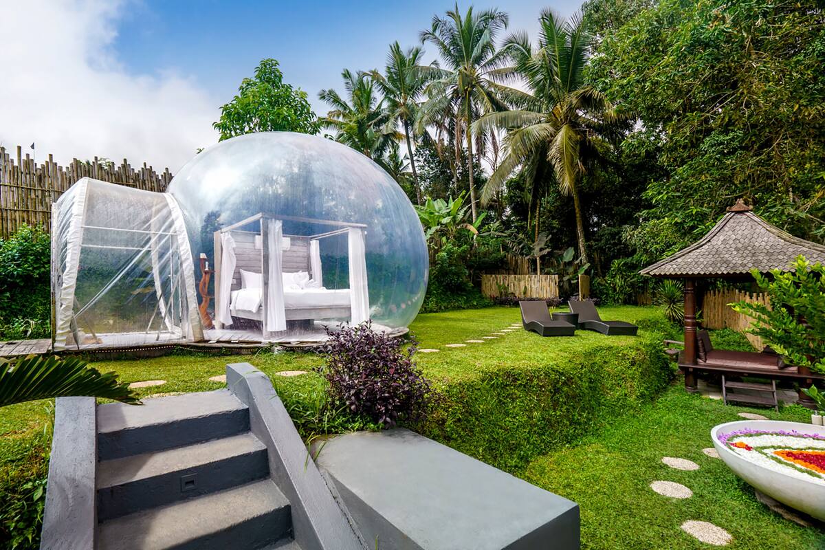 Une structure en forme de dôme transparent abrite un lit, et est entourée d’un jardin luxuriant agrémenté de palmiers et d’un petit kiosque.