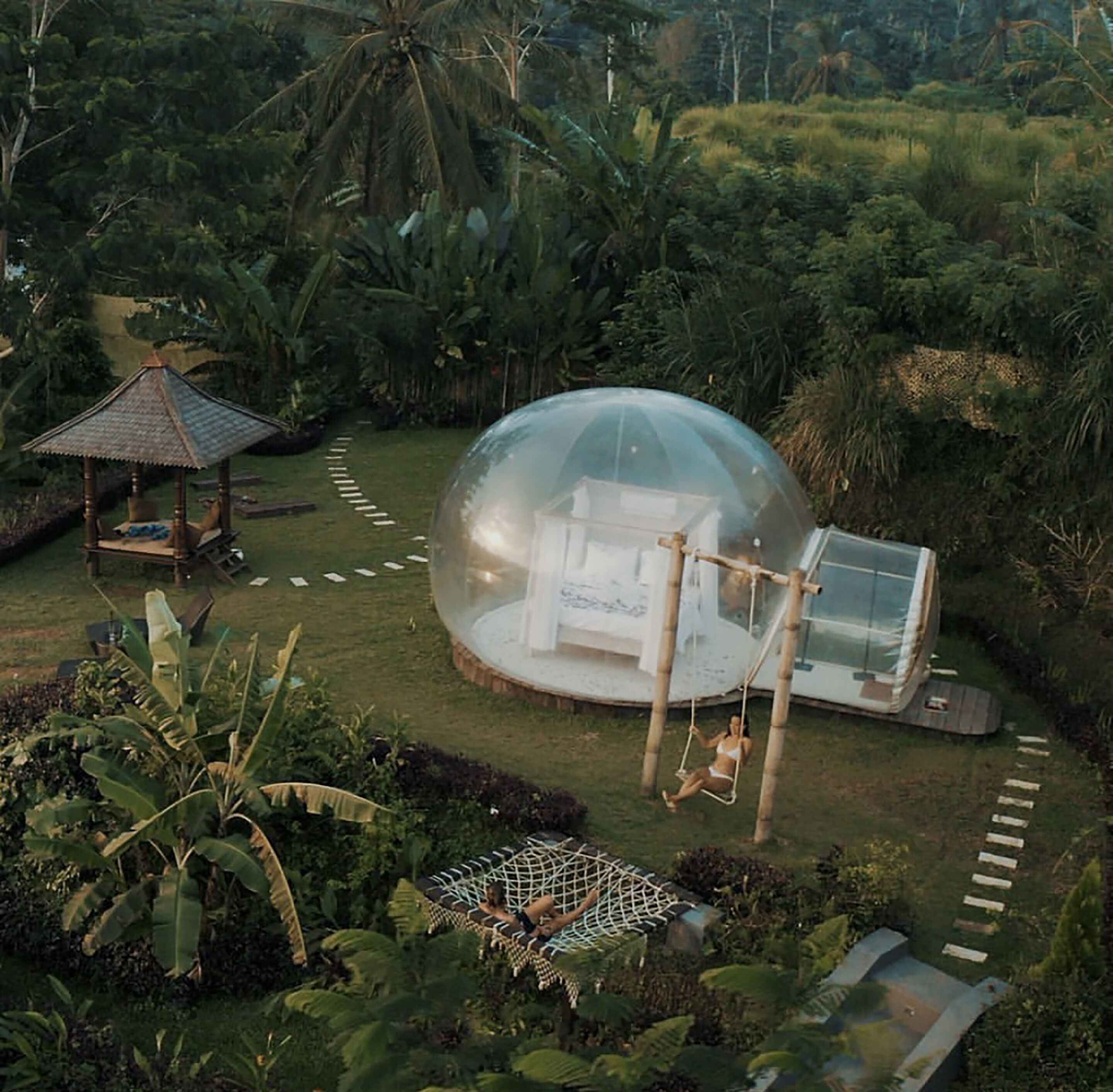 A transparent dome structure with a hammock and a small pavilion in a lush garden.