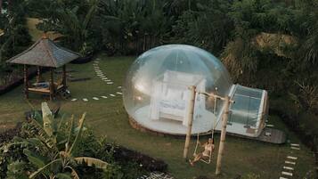 A transparent dome structure with a hammock and a small pavilion in a lush garden.
