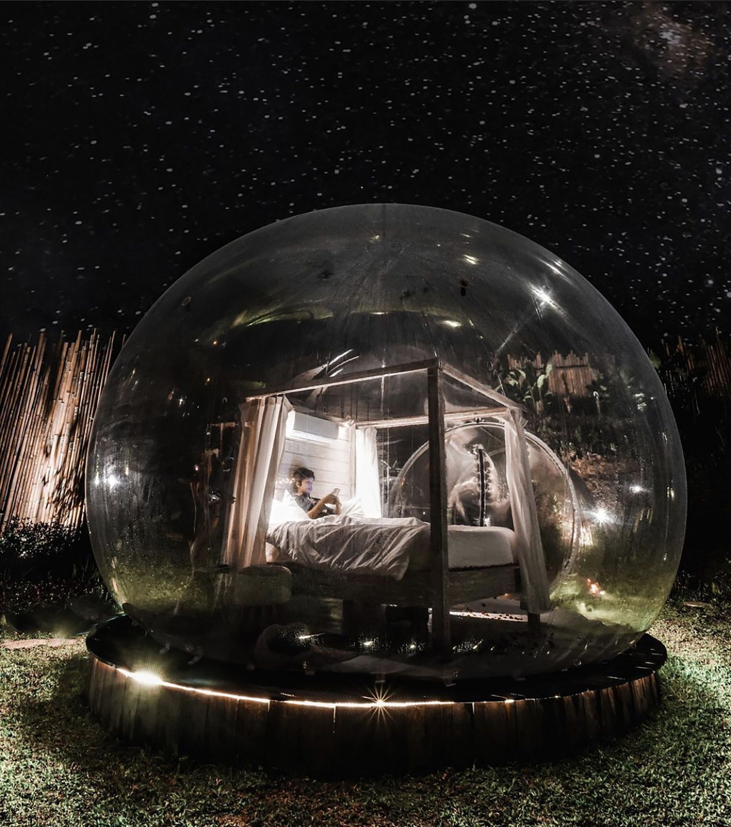 Un dôme transparent abritant un lit et une personne qui lit, le tout entouré d’une clôture en bois et surplombé par un ciel étoilé.