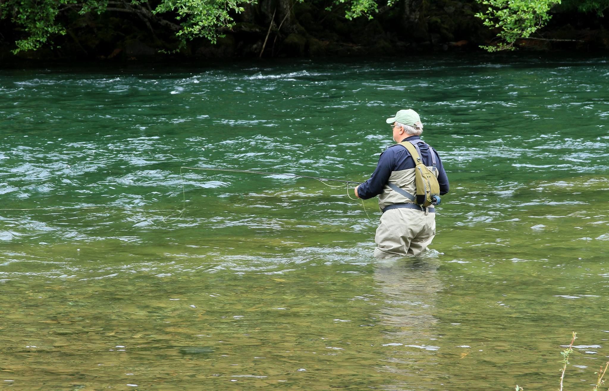 A man standing in a river, wearing a hat and carrying a backpack.