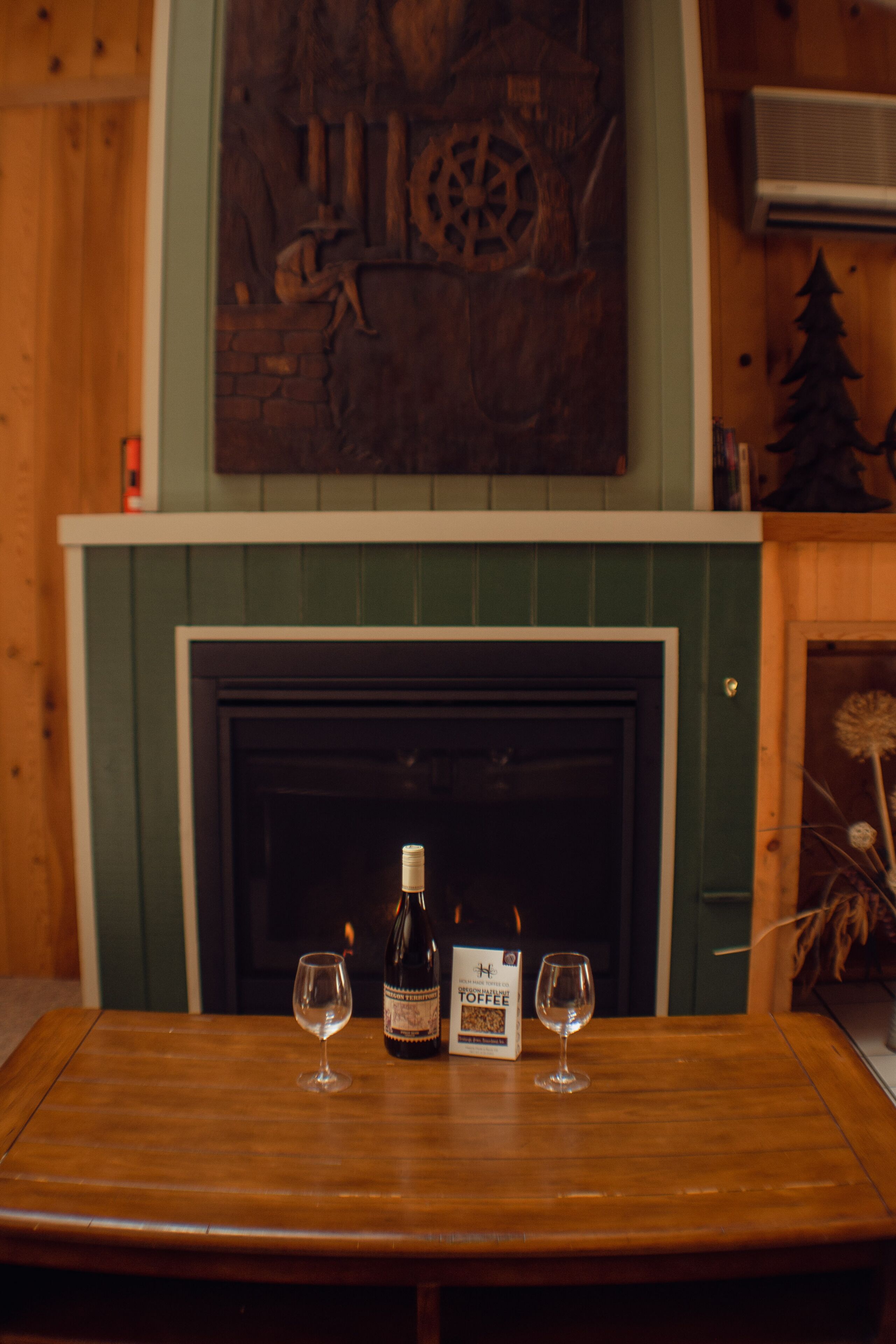 A wooden table with two wine glasses and a bottle of wine, set against a backdrop of a fireplace and a decorative wall piece.