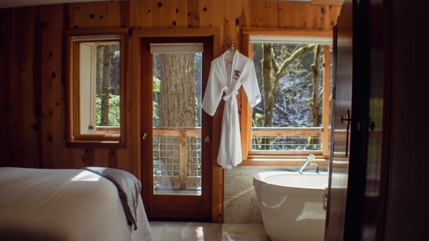 A wooden cabin bedroom with a large bed, a bathroom with a bathtub, and a robe hanging by the door.