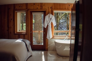 Un dormitorio de cabaña de madera con una cama grande, un baño con bañera y una bata colgada cerca de la puerta.