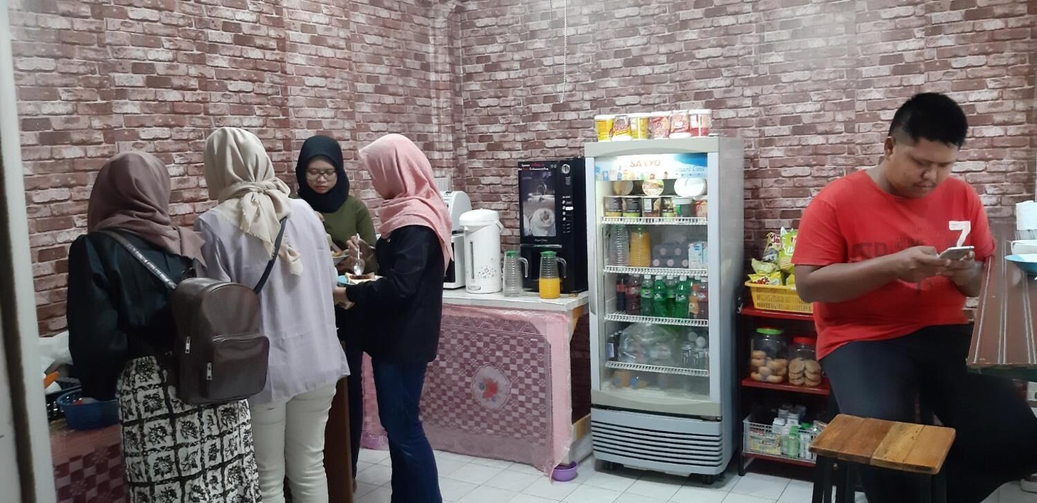 A brick-walled room with a refrigerator stocked with beverages, a table with a pink tablecloth, and a person using a mobile device.