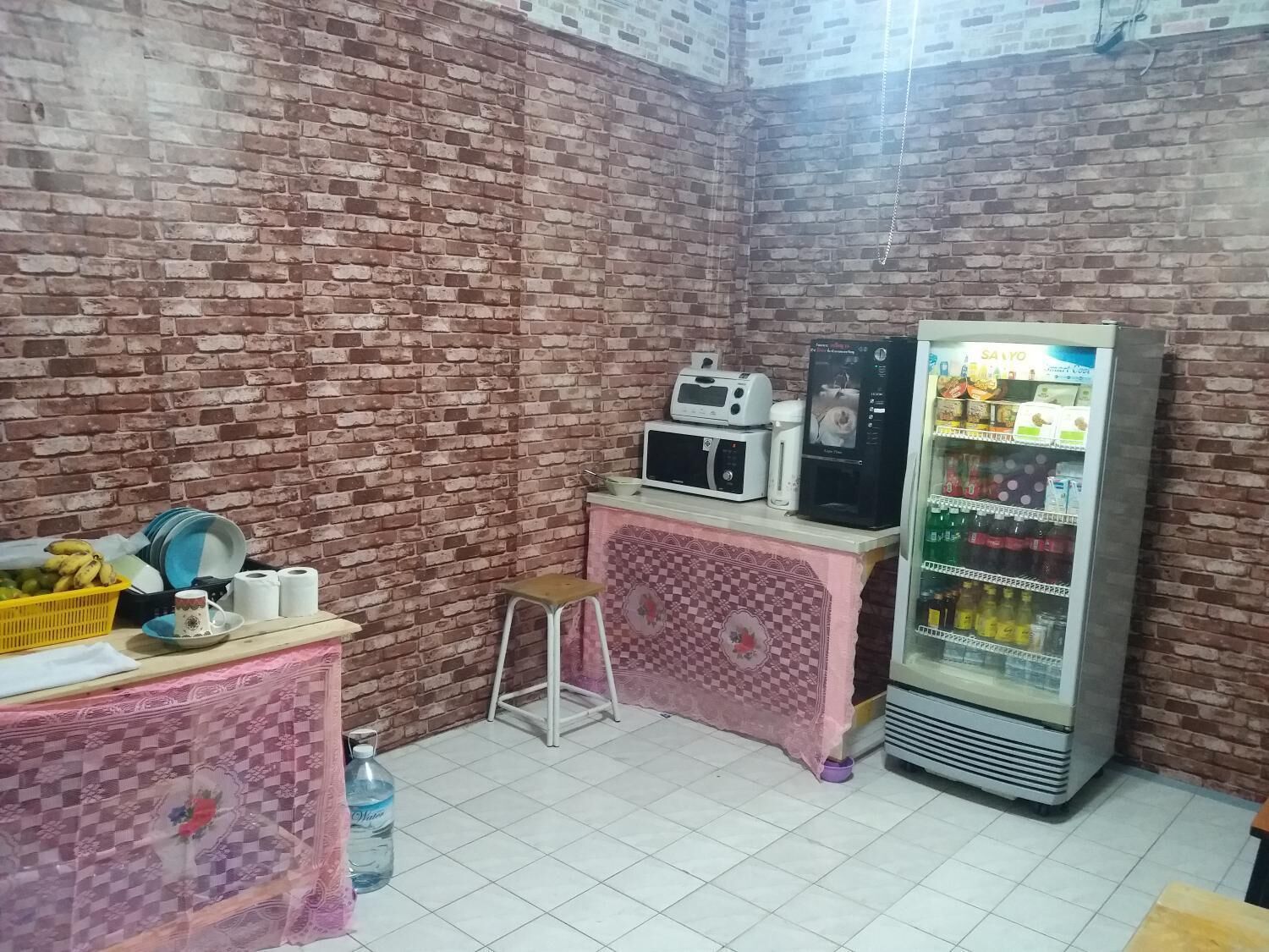 A small room with a brick wall, a microwave, a refrigerator, a small stool, and a table with a basket of bananas and a cup.