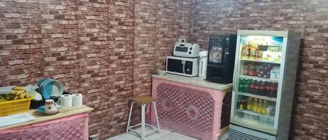 A small room with a brick wall, a microwave, a refrigerator, a small stool, and a table with a basket of bananas and a cup.
