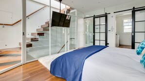 A modern bedroom with a large bed, a TV, a glass door leading to a staircase, and a sliding door.