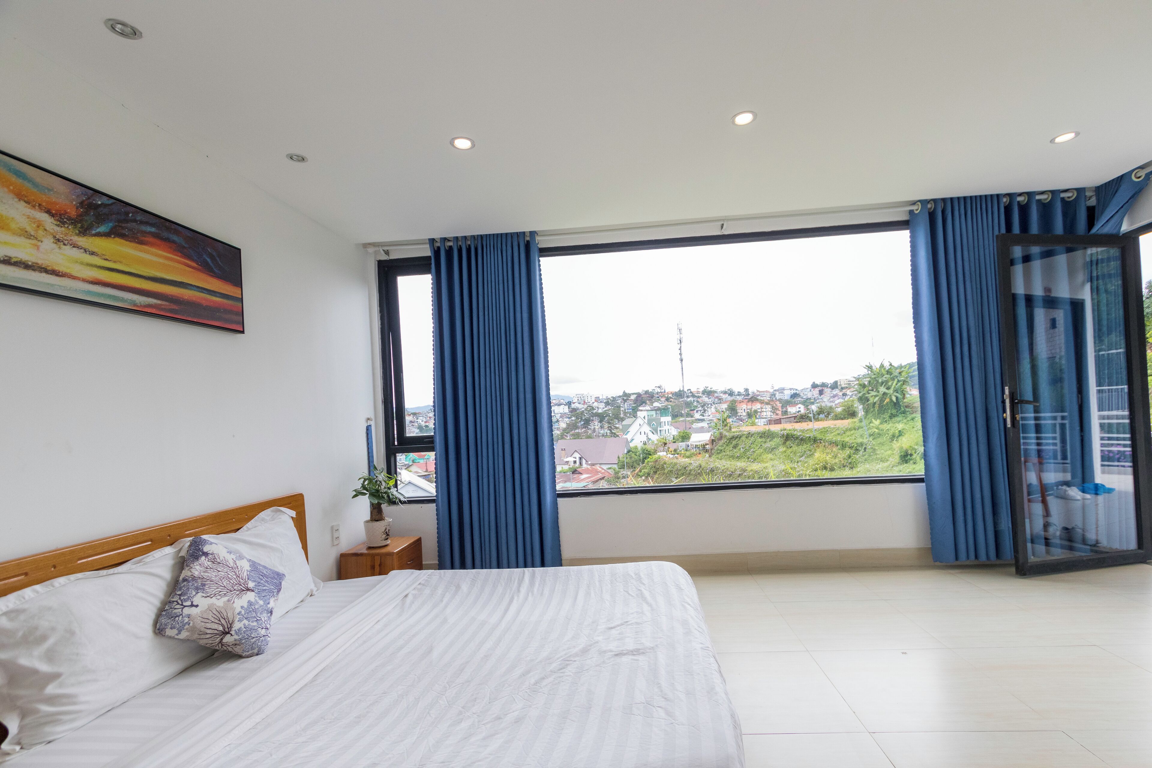 A bedroom with a bed, a window with curtains, a painting on the wall, and a view of the outside.