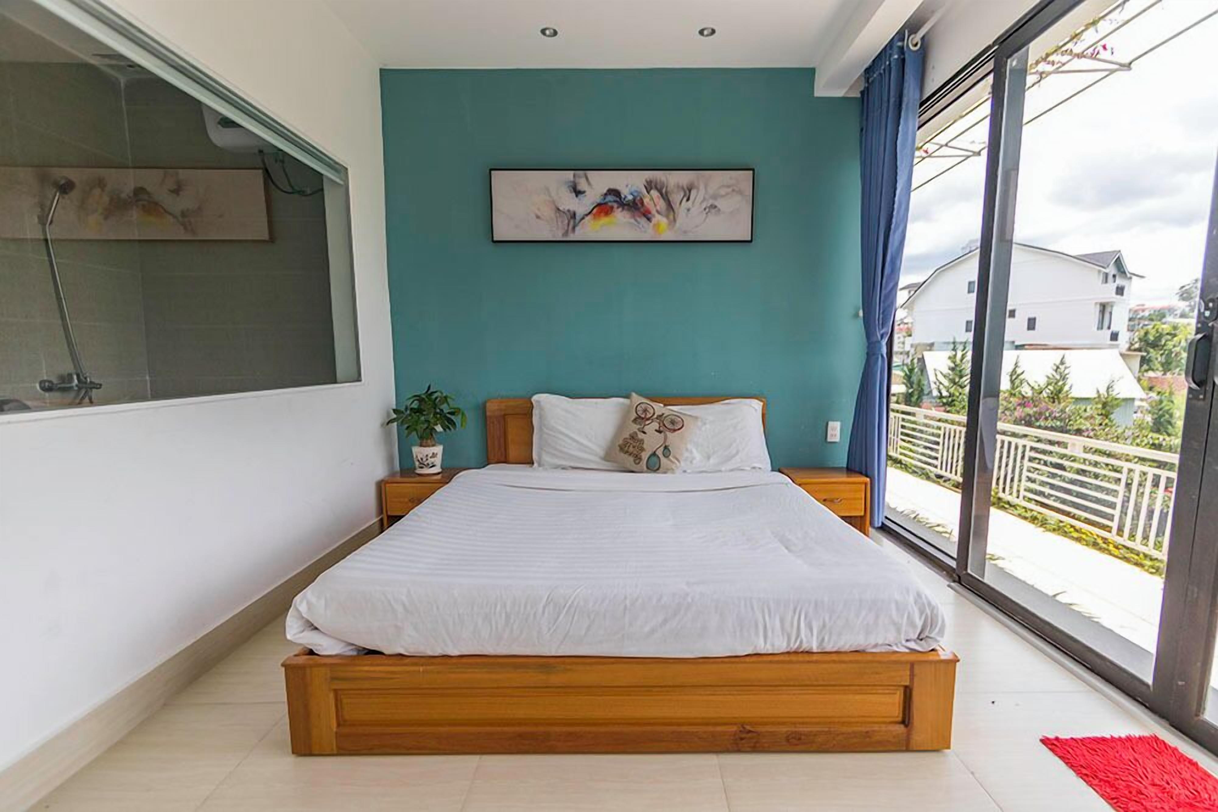 A bedroom with a large bed, a window with a view of the outside, a painting on the wall, and a plant on the nightstand.
