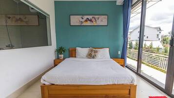 A bedroom with a large bed, a window with a view of the outside, a painting on the wall, and a plant on the nightstand.