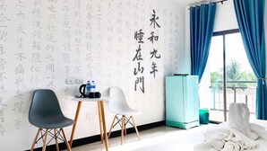 A hotel room with a large bed, two chairs, a small table with a kettle and water bottles, a minibar, and a view of greenery through the window. - Shanmen Art House (Patong)