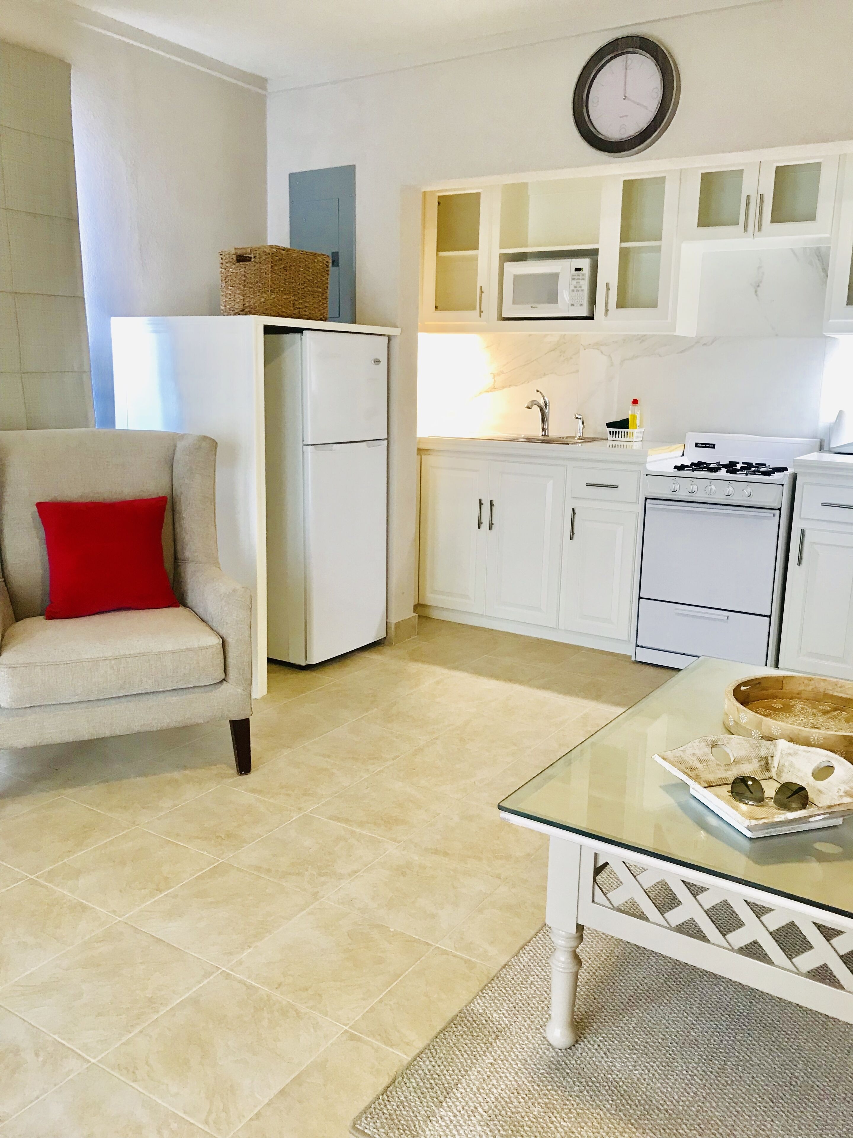A modern kitchen with white cabinets, a refrigerator, a microwave, and a stove. There is a glass table with a tray and a red cushion on a chair.