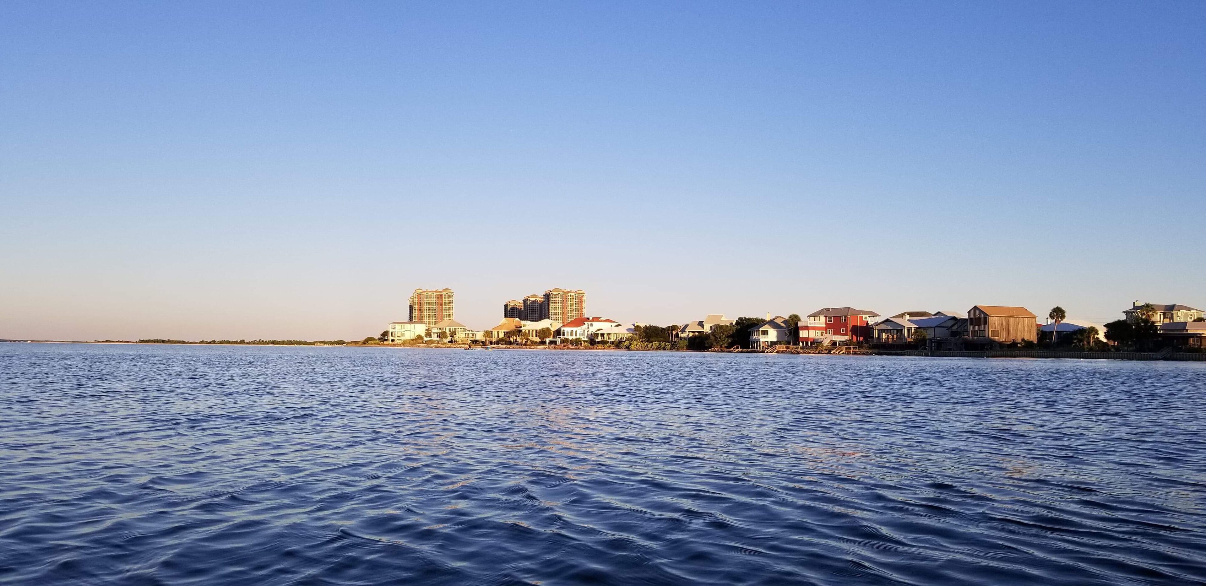 A waterfront cityscape with buildings along the shore, a calm sea, and a clear sky.