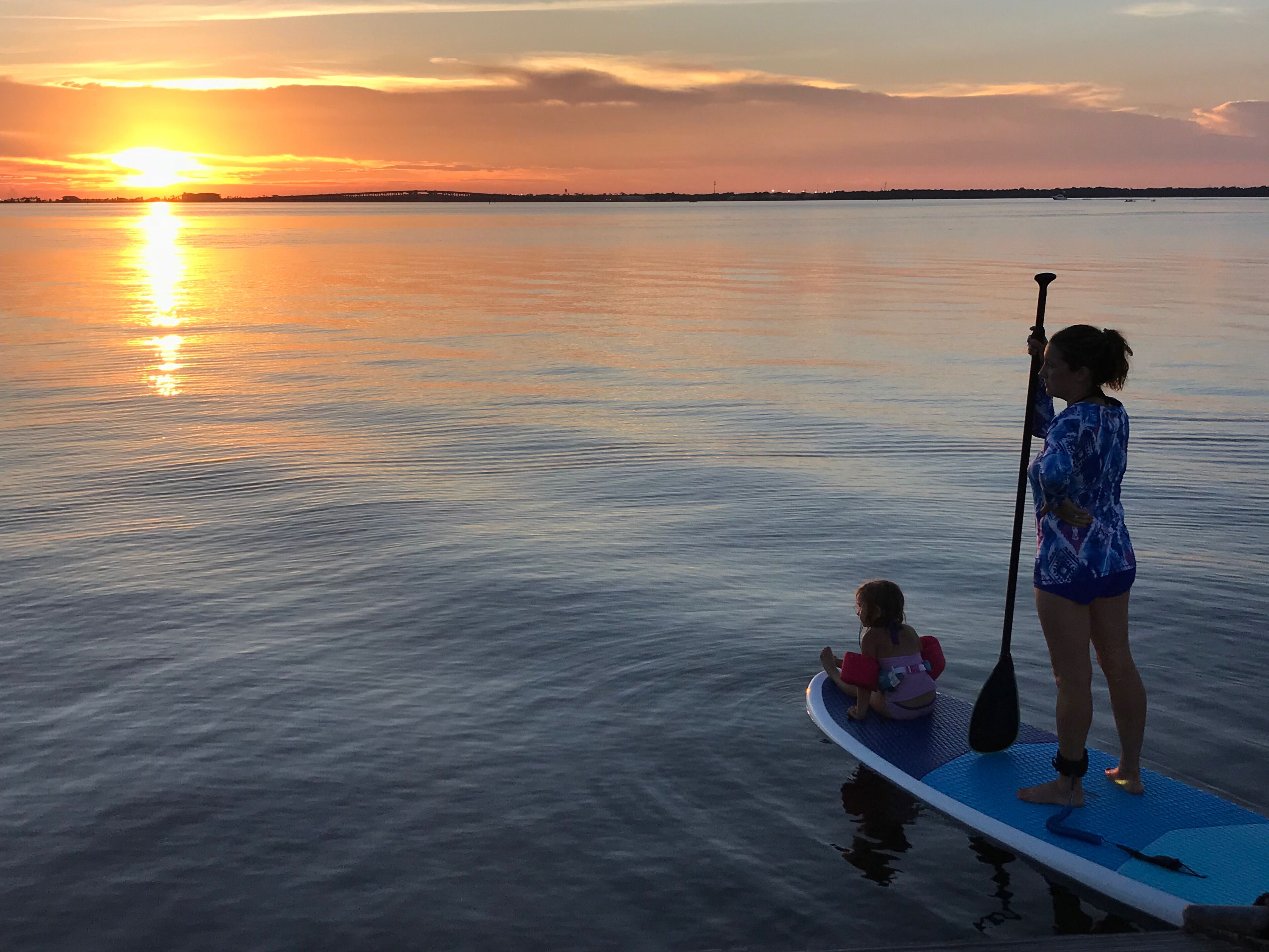 Un atardecer sobre aguas tranquilas, con una tabla de remo y un niño/una niña en ella.