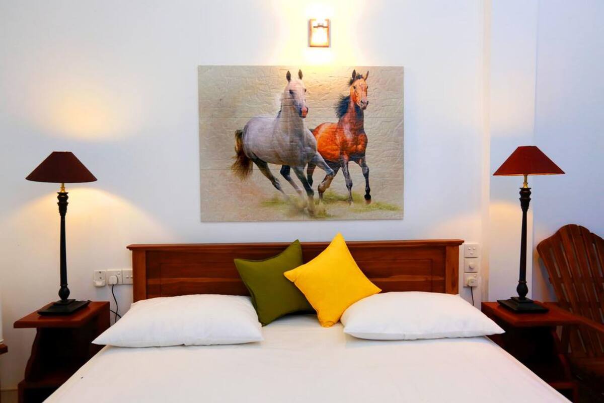 Habitación de hotel con cama, dos mesitas de noche, dos lámparas y un cuadro de caballos en la pared.