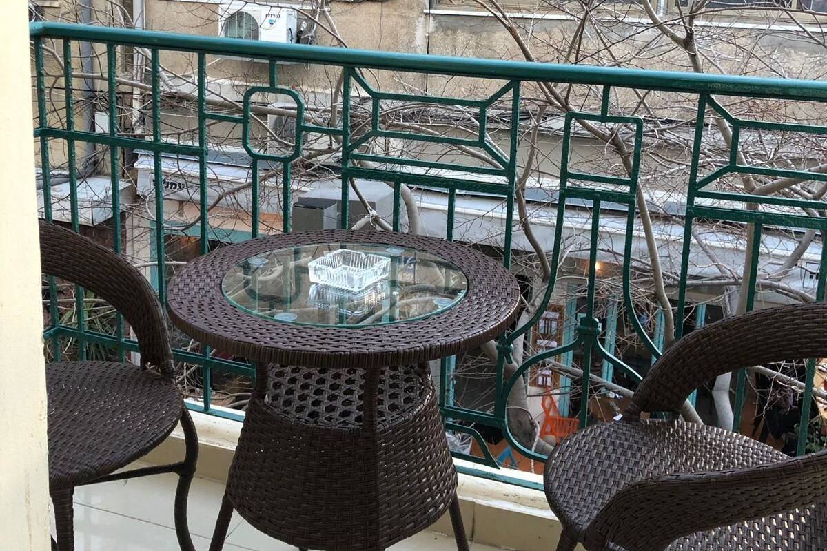 A balcony with wicker furniture and a glass table, overlooking a building with bare trees and a window.