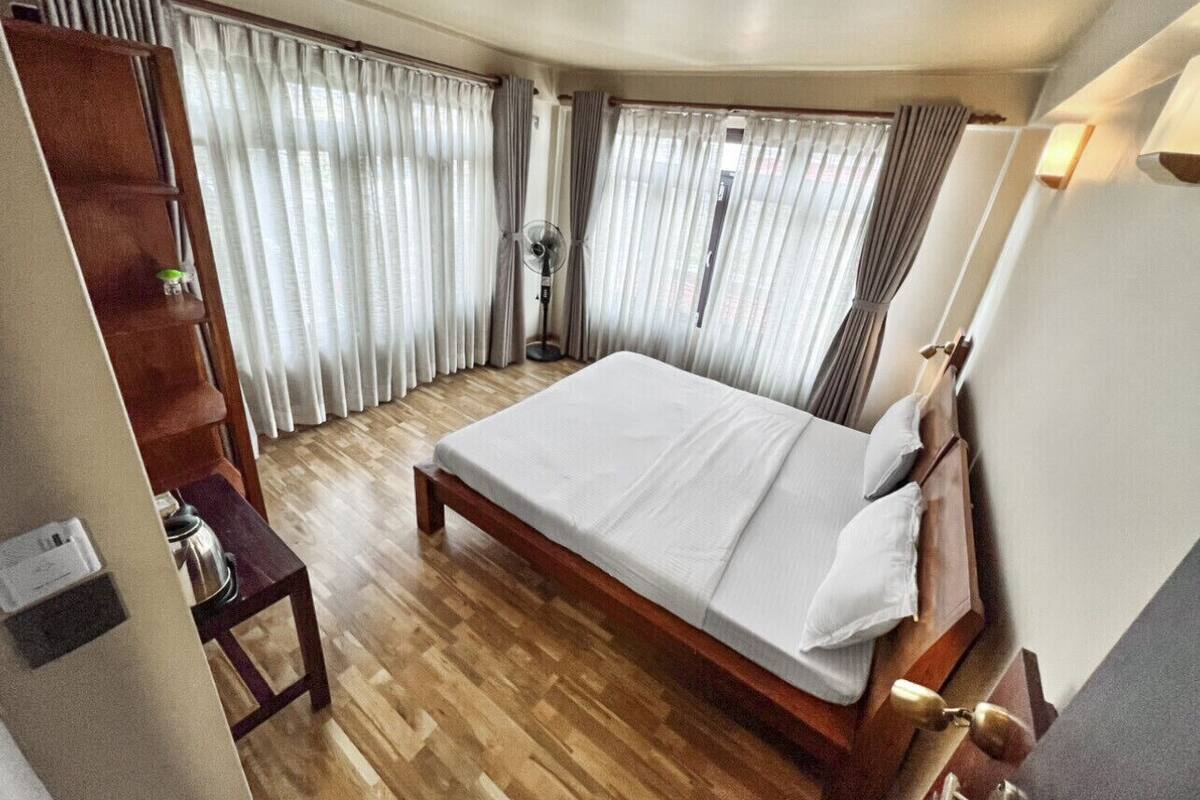 Une chambre d’hôtel avec un lit en bois, un bureau et une chaise. Il y a une fenêtre avec des rideaux et une lampe de sol.