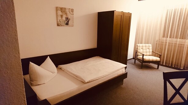 A single bed with a white mattress and pillows, a dark headboard, a wooden wardrobe, a plaid chair, and a window with sheer curtains. - H&S Hotel Wildpferd (Duelmen)
