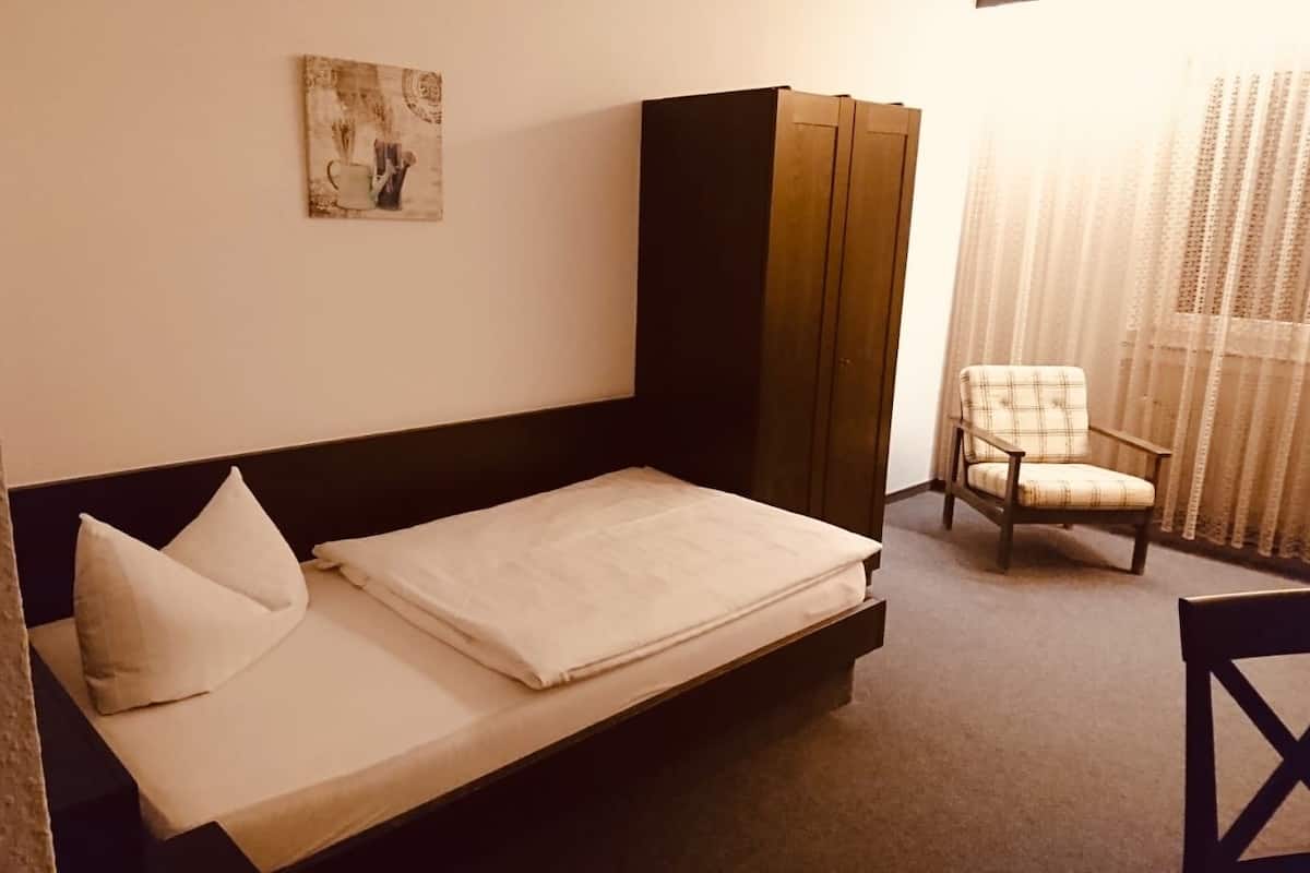 A single bed with a white mattress and pillows, a dark headboard, a wooden wardrobe, a plaid chair, and a window with sheer curtains.