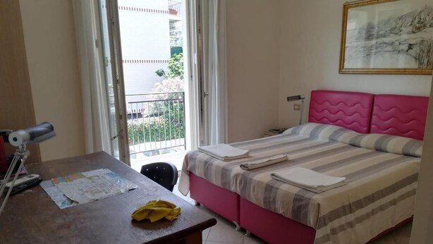 A bedroom with a bed, a desk with a lamp, and a window with a view of a building and greenery. - Anania (Cagliari)