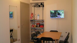 Un comedor con una mesa y sillas de madera, un televisor montado en la pared y una estantería con utensilios de cocina.