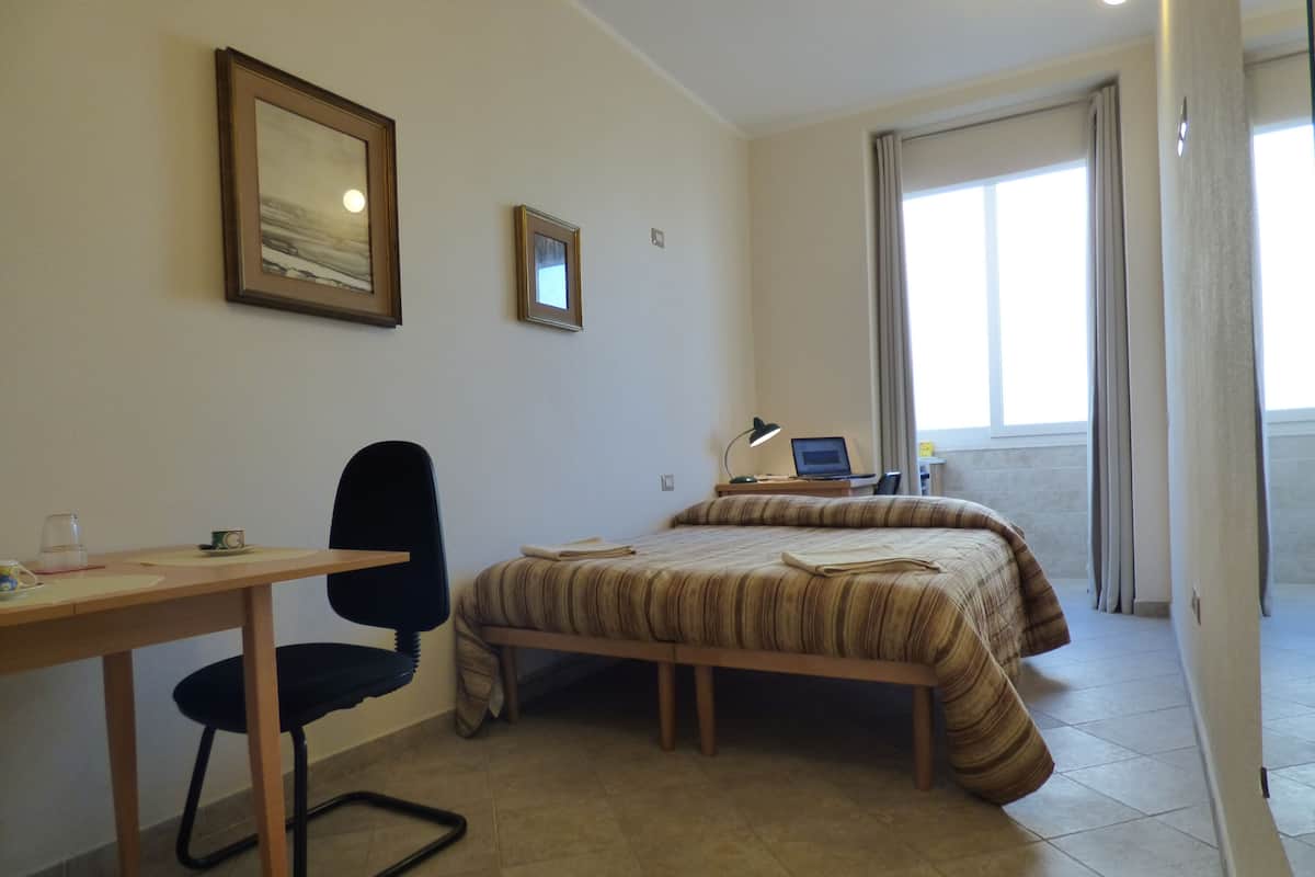 Hotellihuone, jossa on sänky, työpöytä tuolilla, kannettava tietokone ja ikkuna, jossa on verhot.