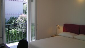 A bedroom with a bed, a nightstand, a lamp, and a sliding glass door leading to a balcony with a view of a building and greenery. - Anania (Cagliari)