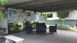 An outdoor patio area with a covered seating area, a white bench, and a bulletin board with various notices. - Ingham Tourist Park (Ingham)