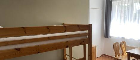 A dormitory room with a bunk bed, a small table, chairs, and a window with curtains.