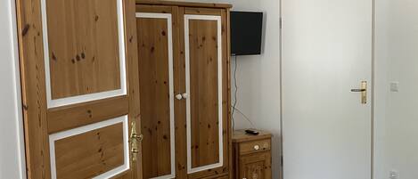 A bedroom with a wooden wardrobe, a bedside table, a bed, and a television mounted on the wall.