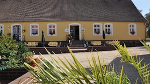 Een geel gebouw met een pannendak, een voordeur en een klein terras met tafels en stoelen.
