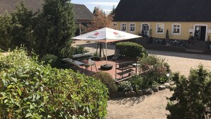 Een terras met tafels en stoelen, een grote parasol, en een gebouw met een gele gevel op de achtergrond.