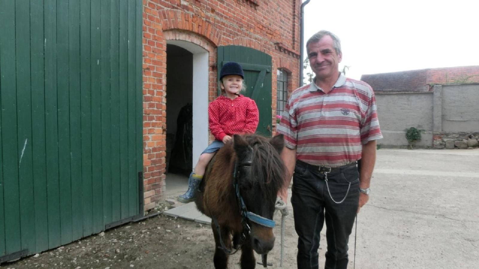 Eine Person steht neben einem Pferd, auf dessen Rücken ein Kind sitzt, vor einem Backsteinhaus mit grüner Tür.