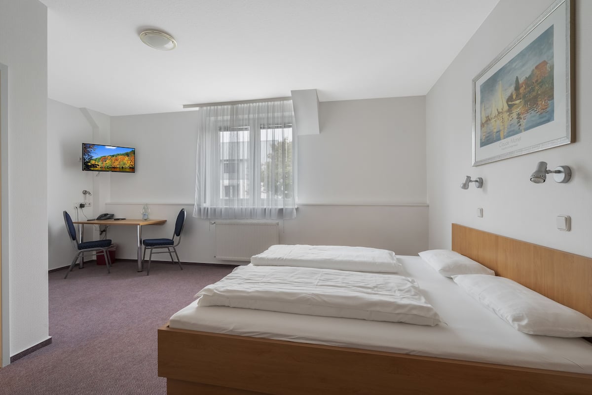 A hotel room with a bed, a desk with a chair, a television, and a window with sheer curtains.