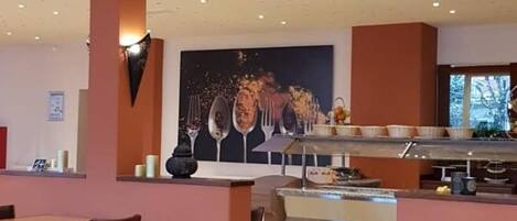 A restaurant with a modern interior, featuring a central counter with a large painting, multiple seating areas with tables and chairs, and a ceiling with recessed lighting.