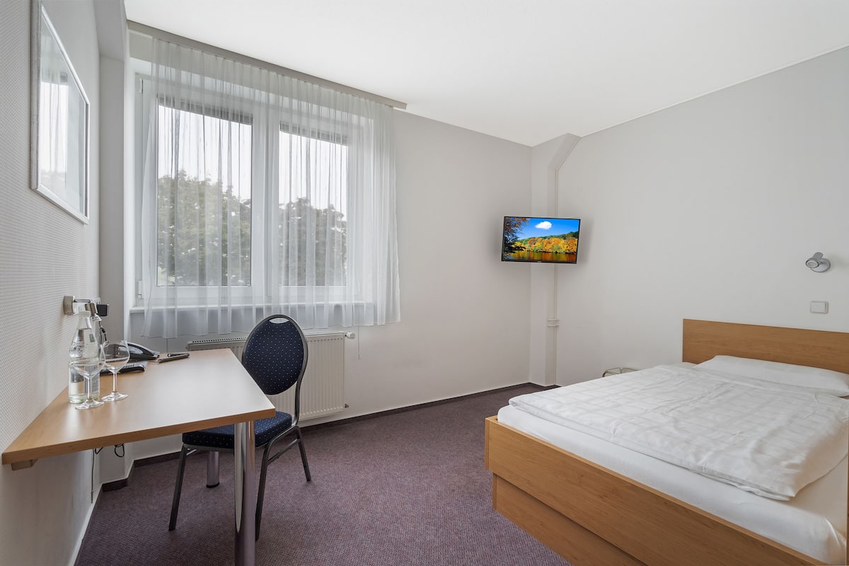 A hotel room with a bed, a desk with a chair, a television, and a window with sheer curtains.