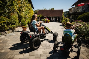 Two children riding toy cars on a paved path with potted plants and a building in the background.