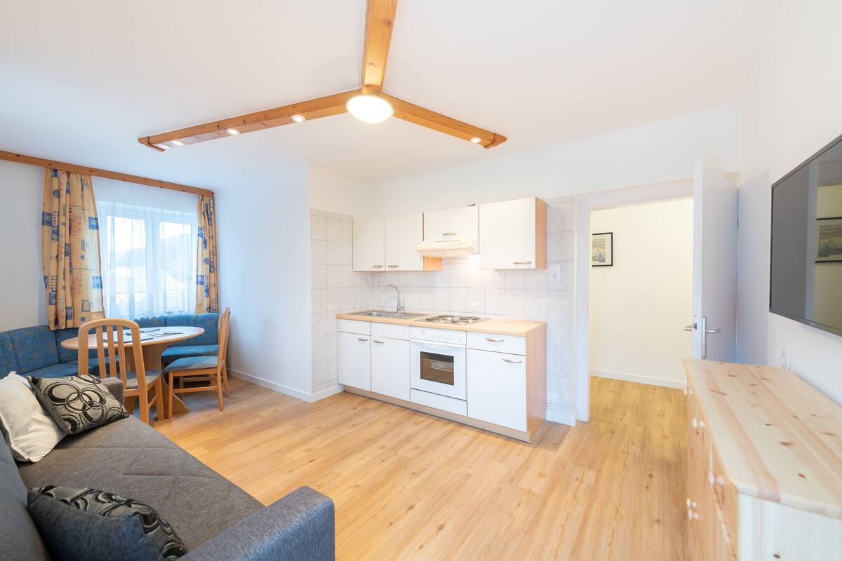 En kompakt opholdsstue med tekøkken, spiseplads med rundt bord og stole, samt en sofa.