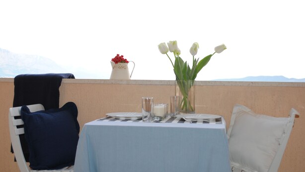 Une table dressée pour deux personnes, avec une nappe bleu clair, des chaises blanches, un vase de fleurs blanches, un pichet de fleurs rouges et un verre de lait.
