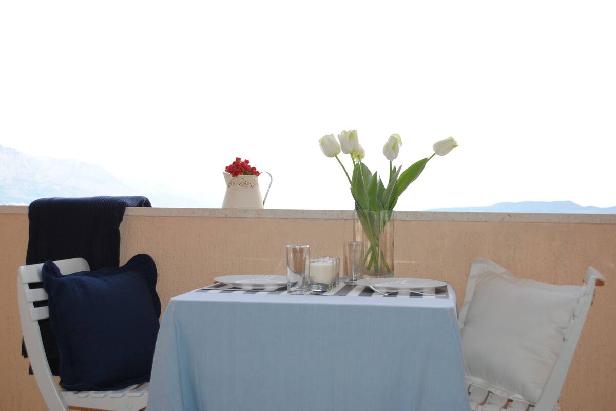A table set for two with a light blue tablecloth, white chairs, a vase with white flowers, a pitcher with red flowers, and a glass of milk.