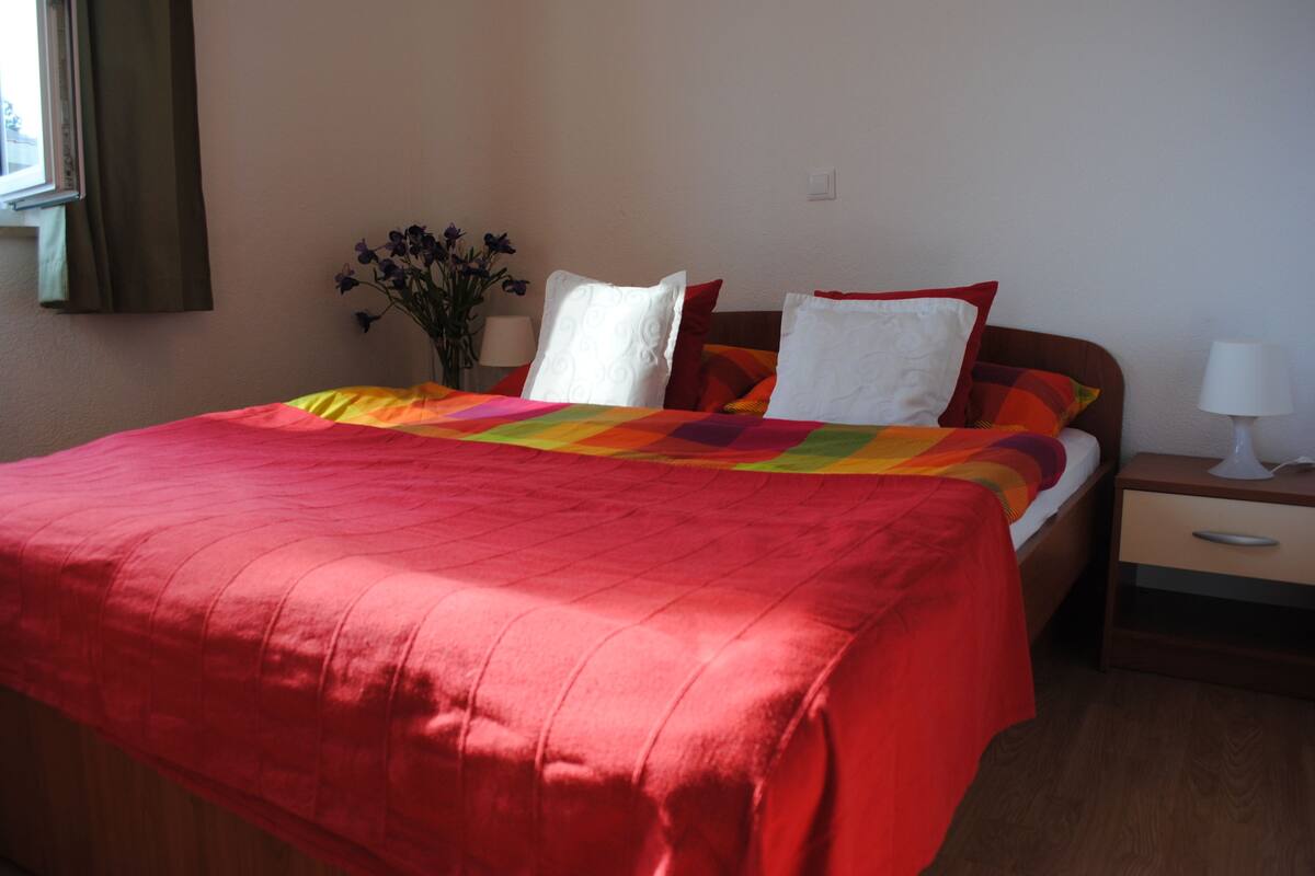 A neatly made bed with a red blanket and pillows, a bedside table with a lamp, and a vase with flowers on the nightstand.