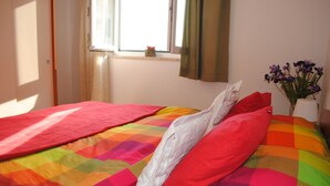A bed with a colorful quilt, a window with curtains, and a vase with flowers. - Mime 2 (Omis)