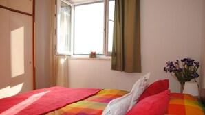 A bedroom with a colorful quilted bedspread, a window with curtains, and a vase with flowers on the windowsill. - Mime 2 (Omis)