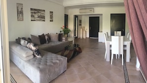 Ein Wohnzimmer mit einem grauen Ecksofa, einem Glastisch und einem Essbereich mit weißen Stühlen und einem Tisch.